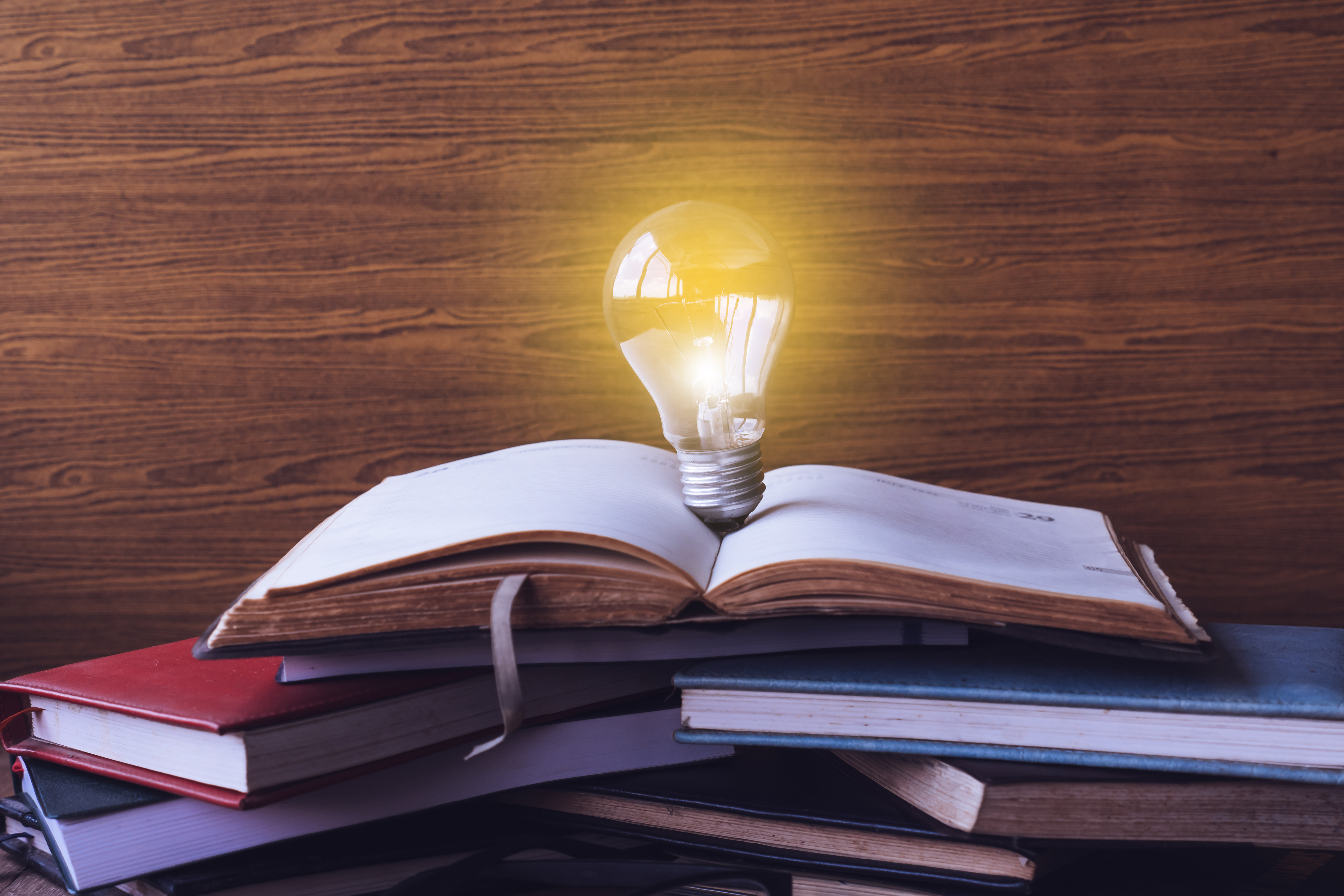 Open book with light bulb and hardback books on wood wall background.